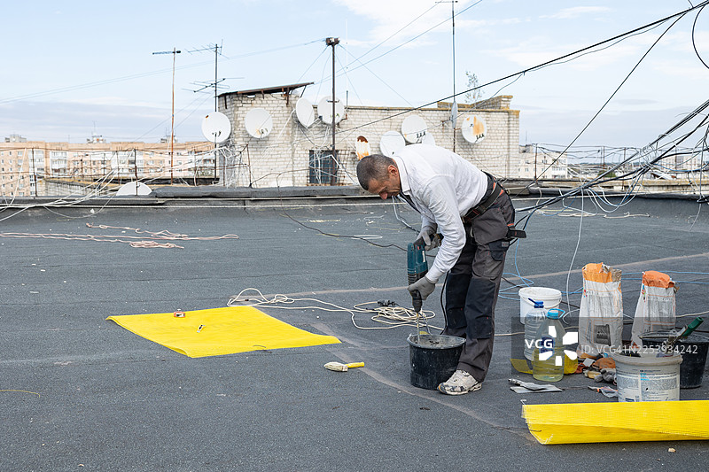 建筑工人在屋顶上用电钻搅拌桶里的砂浆。图片素材