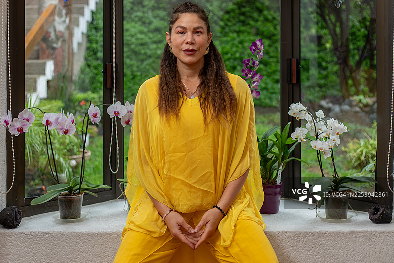 中年女性坐在窗边，双手交叠，周围环绕着兰花，寻求内心的平静与健康。图片素材