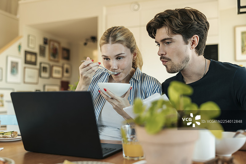 年轻女子在家一边吃早餐，一边和男友用笔记本电脑规划预算图片素材