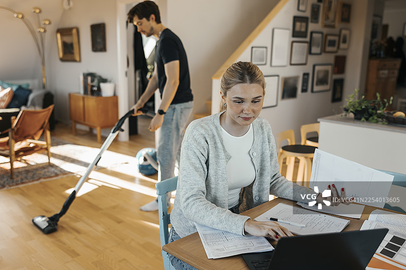 俯视视角，年轻的女自由职业者在家中进行会计工作，男友在背景中打扫地板图片素材