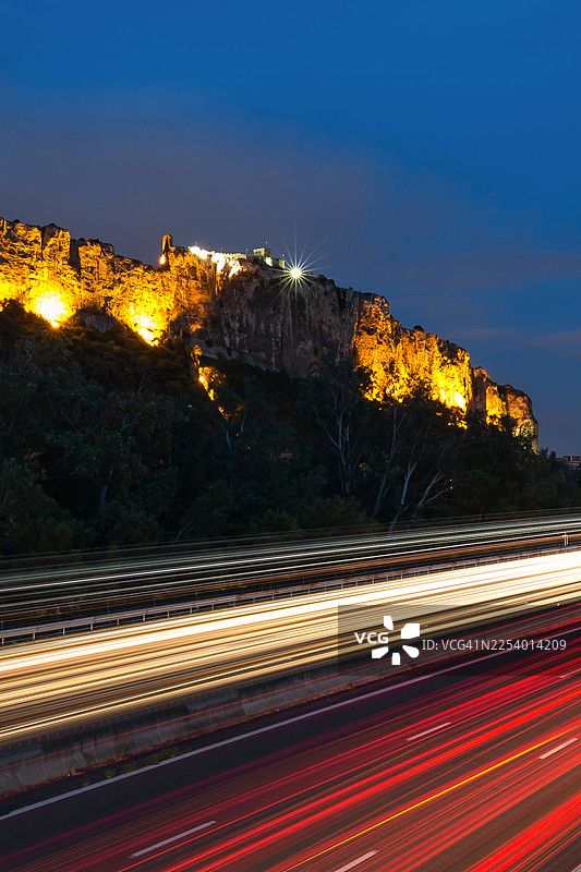 蓝色时分，高速公路上车灯闪耀，背景中有一座灯火辉煌的历史悠久的堡垒图片素材