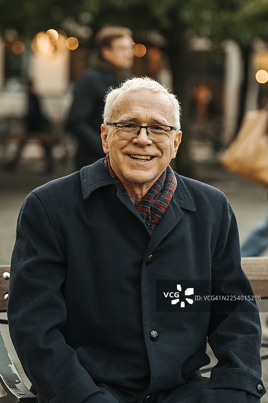 一位穿着长外套、面带微笑的老年男子坐在街边长椅上的肖像图片素材
