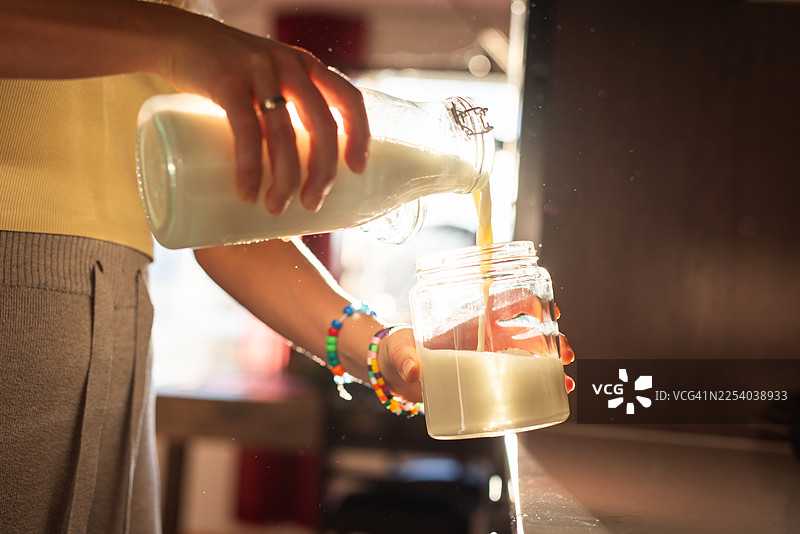 一位未知的女性正在厨房里，在晨光的照耀下，将新鲜牛奶从玻璃瓶倒入罐中。图片素材