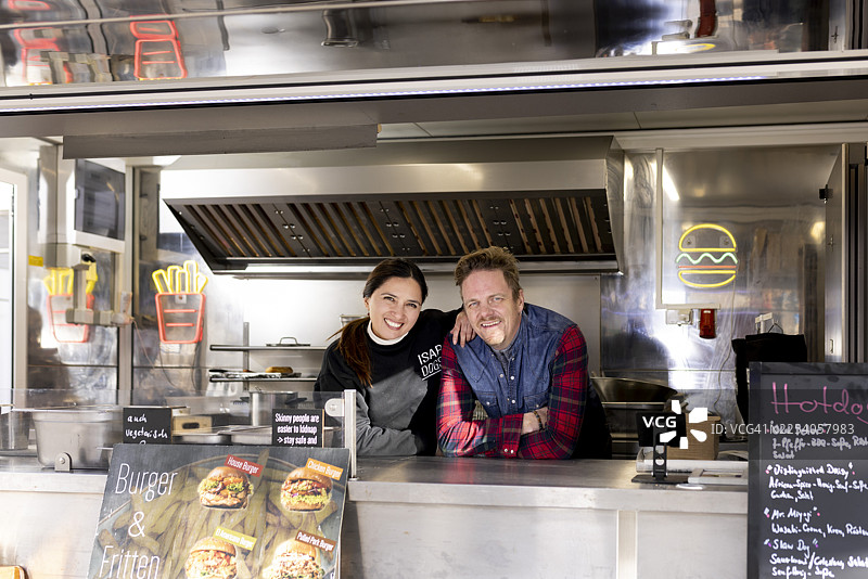 一对经营街头食品卡车的成熟夫妇/商业伙伴，倚靠在他们其中一辆食品卡车的厨房柜台上。图片素材