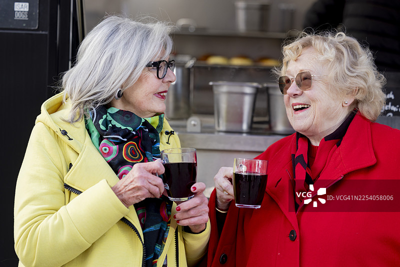 两位快乐、美丽、充满活力的老年女性在寒冷的天气里，在餐车前享用她们点的热红酒（Glühwein）。图片素材