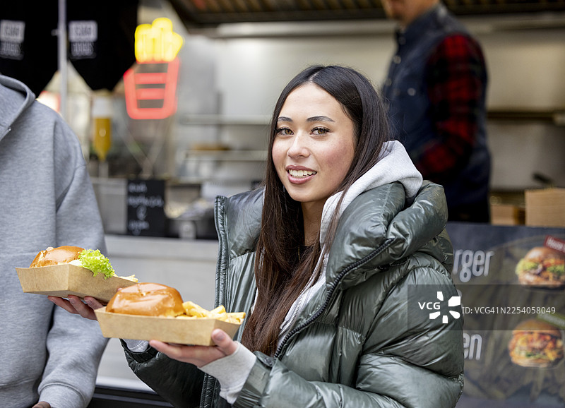 一位有着长棕发的混血女学生在寒冷天气里，在餐车前拿着用纸盒打包好的美食汉堡薯条，准备带走。图片素材