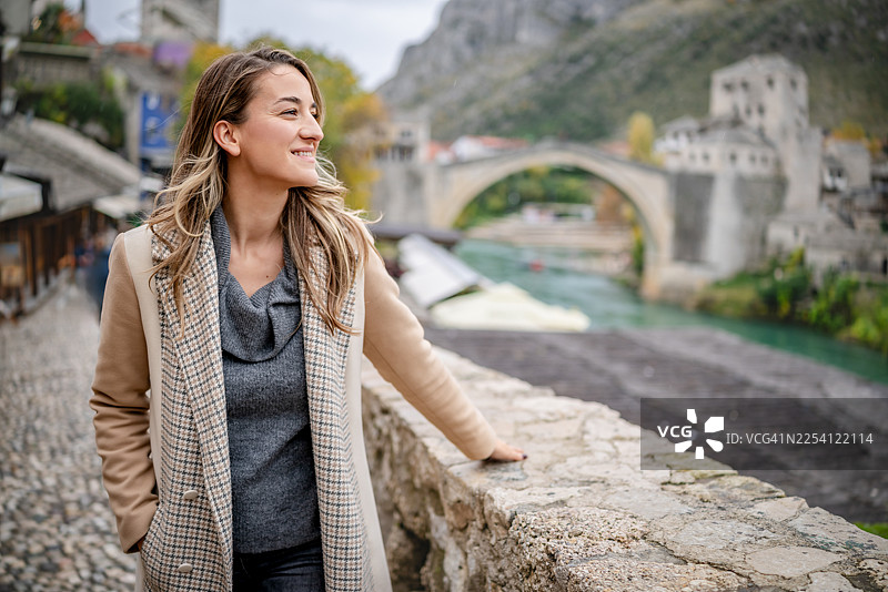 一位年轻女子在探索波黑莫斯塔尔市时，在河边享受片刻宁静图片素材