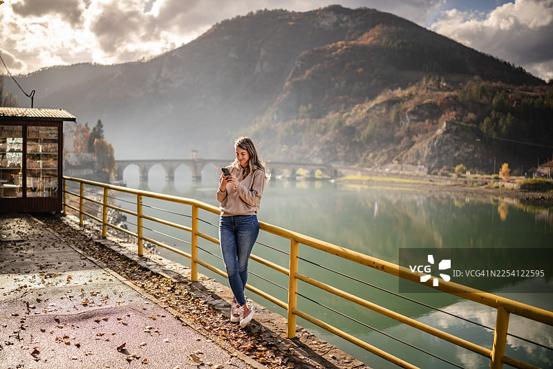 年轻女子在波黑维谢格拉德的水边享受时光，并借机查看手机图片素材