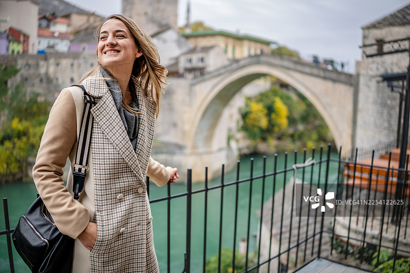 一位年轻女子在多云的日子里，在绿色的水域和历史建筑的环绕下，在莫斯塔尔一座著名桥梁附近享受她的时光图片素材