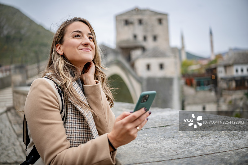 波斯尼亚和黑塞哥维那，阴天，一名年轻女子在莫斯塔尔欣赏风景，同时使用智能手机。图片素材