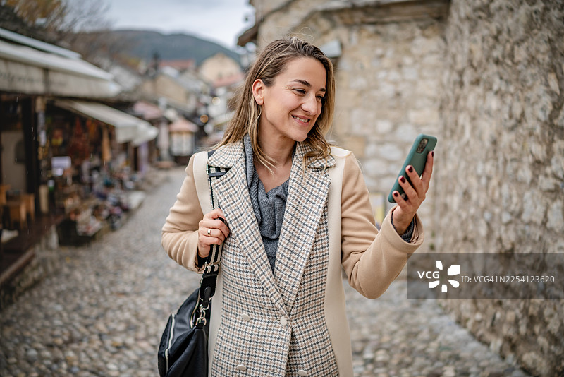 年轻女子走在鹅卵石街道上，用智能手机探索莫斯塔尔图片素材