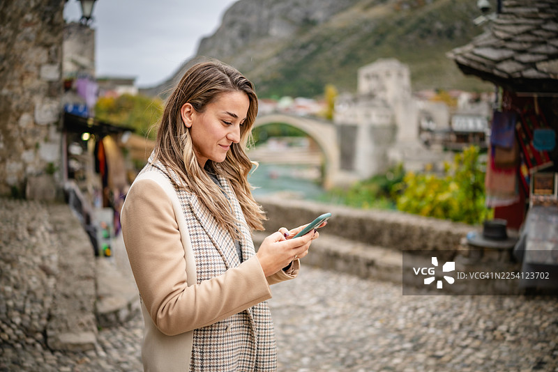 年轻女子在波斯尼亚和黑塞哥维那的莫斯塔尔，于古老河畔户外环境中边使用手机边探索图片素材