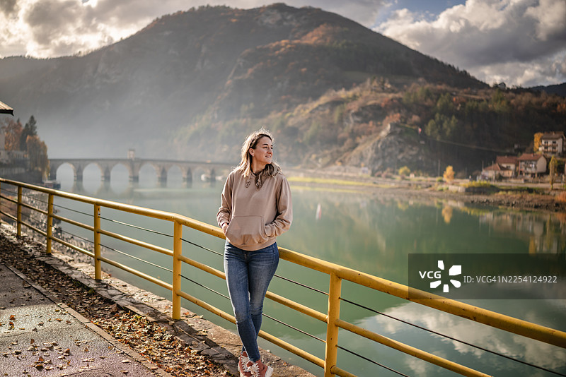 年轻女子在晴朗的日子里站在波黑维谢格拉德的河边，欣赏着群山的美景图片素材