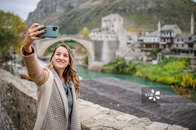 年轻女子在莫斯塔尔河边，一边欣赏户外风景和历史建筑，一边自拍图片素材