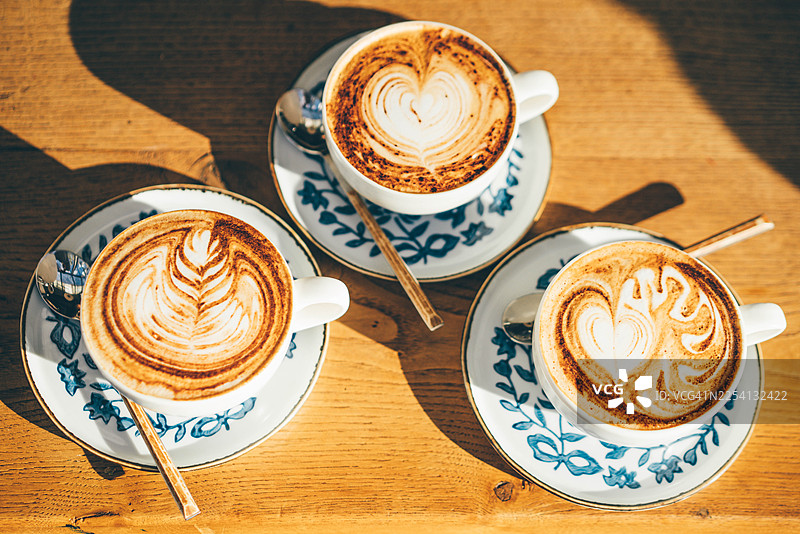 明亮的晨光下，木桌上摆放着三杯卡布奇诺，上面饰有精致的拿铁拉花。这张俯视角度的拍摄捕捉了一个社交连接的瞬间以及咖啡文化的精致之美。图片素材