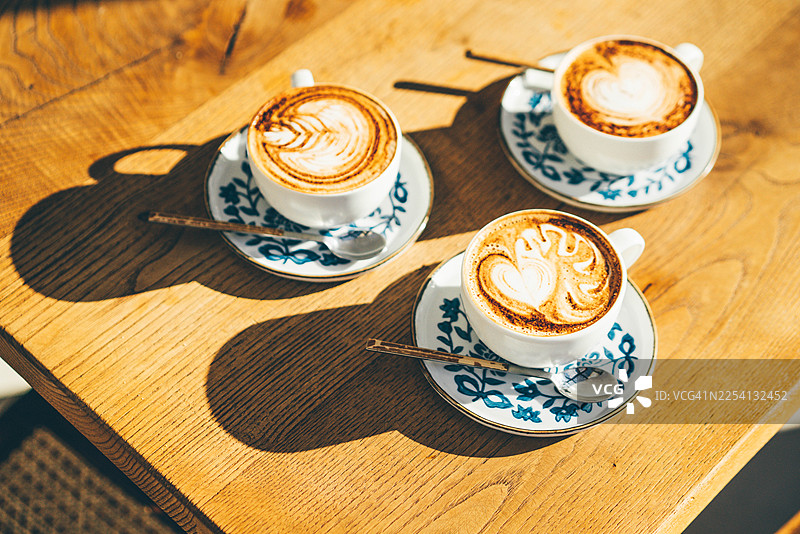 明媚的晨光下，木桌上摆放着三杯卡布奇诺，上面饰有精致的拉花。这张俯拍照片捕捉了社交连接的瞬间以及咖啡文化的精致之美。图片素材