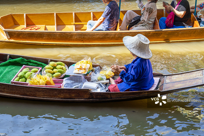 泰国叻丕府丹嫩沙多水上市场，一位商贩在装满新鲜绿色和切片黄色芒果的传统木船上，与狭窄运河里的游客互动图片素材