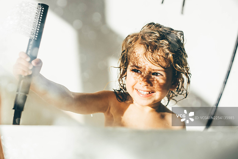 一个卷发、笑容可掬的小女孩，在阳光明媚的浴缸里玩耍，手里拿着淋浴喷头。这张抓拍的肖像捕捉到了快乐的发现、童年的纯真以及日常的健康习惯。图片素材