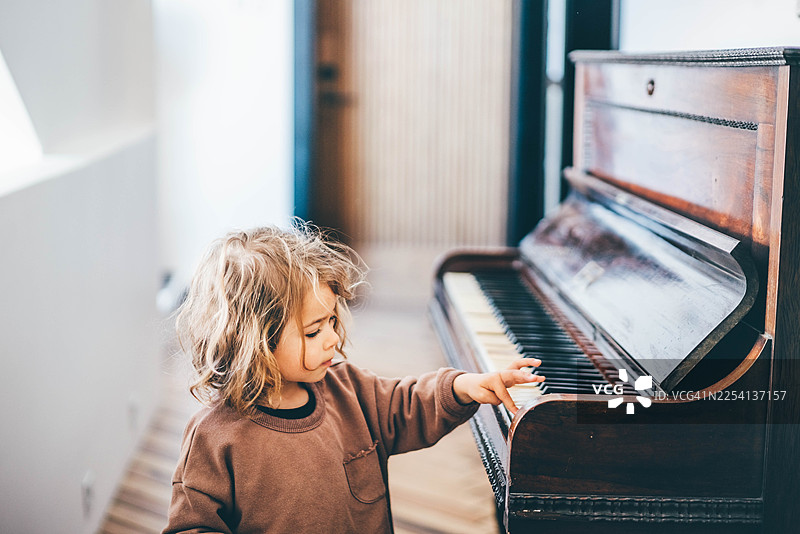 一个金发蓬乱的女孩好奇地伸出手，触碰明亮现代家居中的老式木制钢琴琴键。图片素材