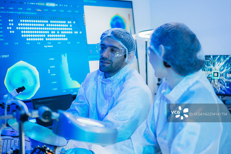 一位身穿无菌防护服的专业技术人员在无尘室中，戴着手套，检查色彩鲜艳的半导体晶圆，象征着先进的微芯片技术和质量控制。图片素材