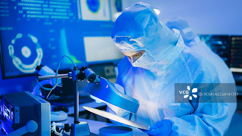 身穿无菌防护服的专业技术人员在半导体制造工厂的洁净室内，戴着手套检查色彩鲜艳的反射性半导体晶圆，代表着先进的微芯片技术和质量控制。图片素材
