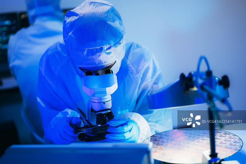 身着无菌防护服的专业技术人员在半导体制造厂的洁净室内，戴着手套检查色彩鲜艳的反射性半导体晶圆，代表着先进的微芯片技术和质量控制。图片素材