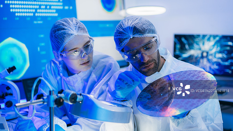 身穿无菌防护服的专业技术人员在半导体制造工厂的洁净室内，戴着手套检查色彩鲜艳的反射性半导体晶圆，代表着先进的微芯片技术和质量控制。图片素材