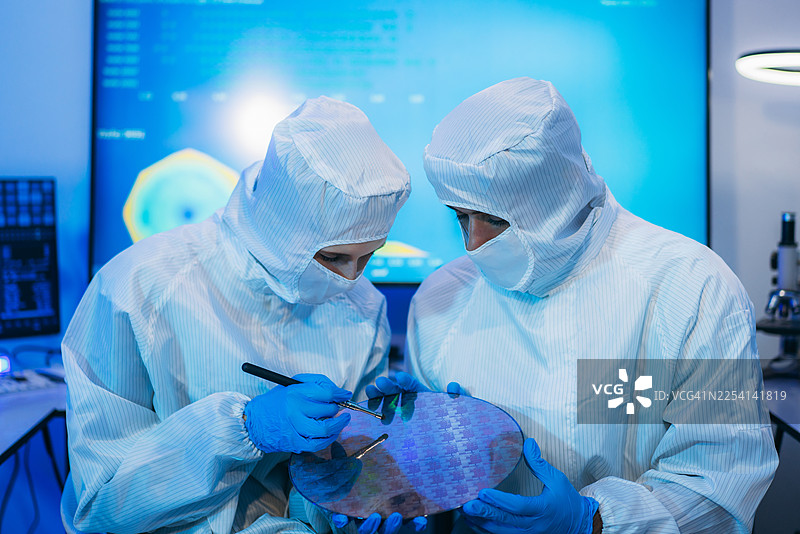 身穿无菌防护服的专业技术人员，在半导体制造工厂的洁净室内，戴着手套检查色彩鲜艳的反射性半导体晶圆，代表着先进的微芯片技术和质量控制。图片素材