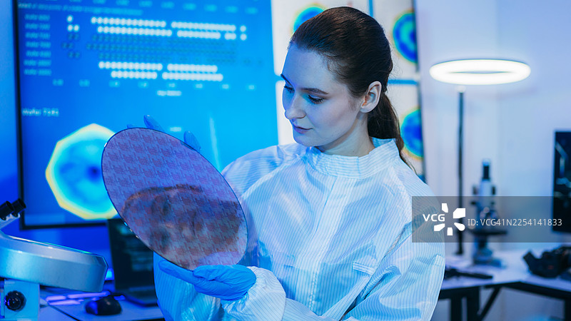 专业技术人员身穿无菌防护服，戴着手套，在半导体制造厂的洁净室内检查色彩鲜艳的反射性半导体晶圆，代表着先进的微芯片技术和质量控制。图片素材