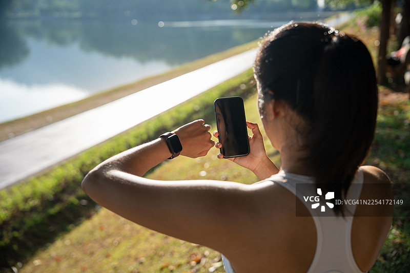 年轻亚洲女性使用智能手表和智能手机上的跑步应用程序来追踪配速和时间。使用可穿戴技术进行锻炼。女性使用智能手机和智能手表。现代科技与健康生活方式的概念。图片素材