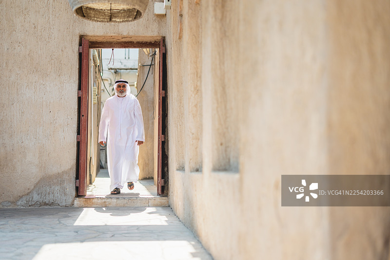 身穿传统白色坎杜拉的阿联酋老年男子，站在阳光明媚的法希迪历史建筑门口图片素材