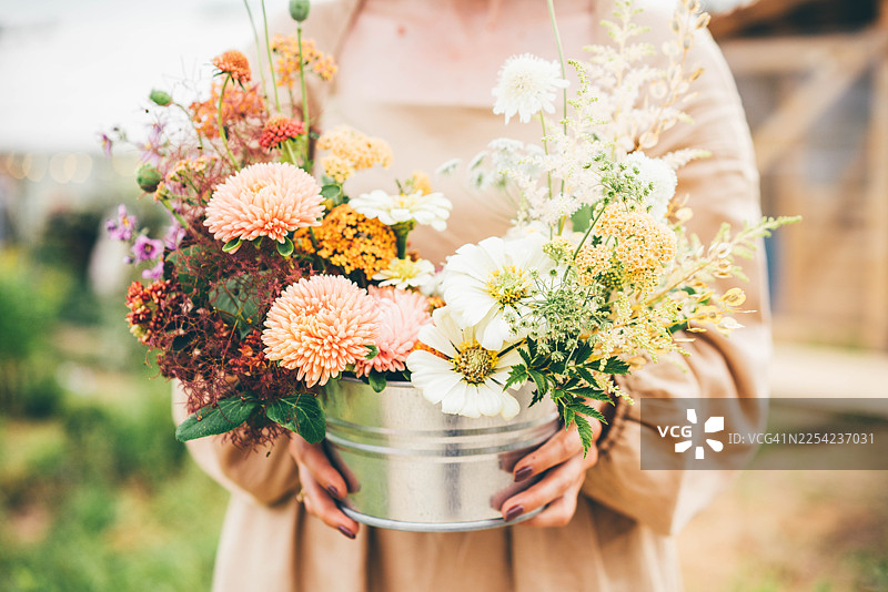 一个人拿着一个乡村风格的金属桶，桶里装满了鲜艳的、手工采摘的秋季花束。柔和的自然色调和手工的插花方式，让人联想到丰收、本地农业和季节性植物的美丽。图片素材