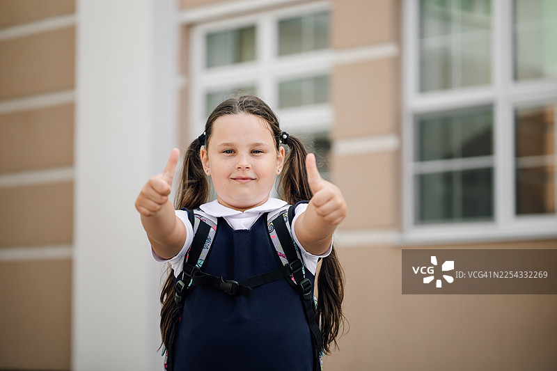 穿着校服的年轻女孩在现代建筑外竖起大拇指，准备好学习和交友图片素材