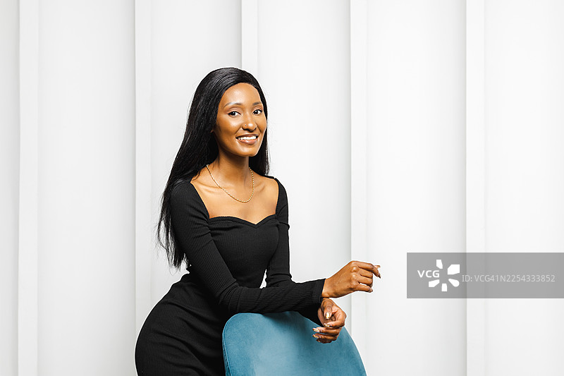 身穿黑色连衣裙的自信女性，在白色背景的影棚中，坐在蓝色椅子上摆姿势图片素材