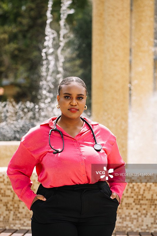 粉色衬衫、戴着听诊器的自信女医生在喷泉旁的户外肖像图片素材