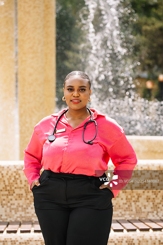 身穿粉色衬衫、佩戴听诊器的自信女医生在喷泉旁的户外肖像图片素材