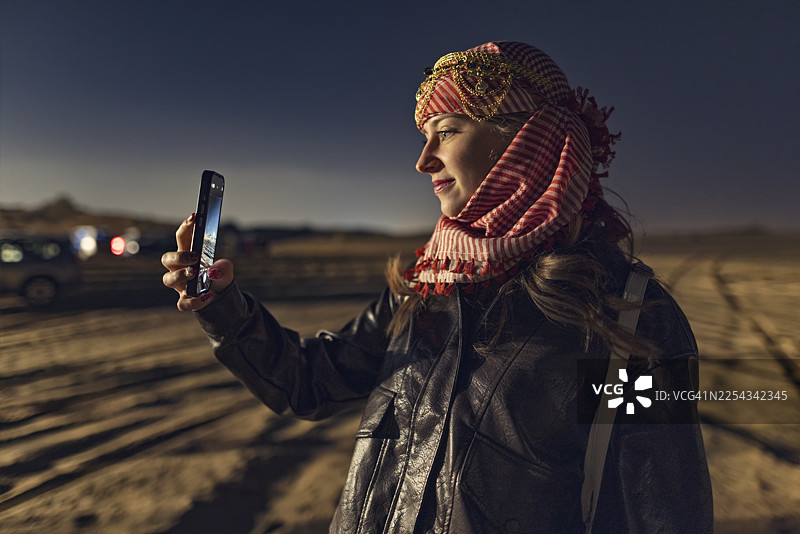 年轻女子游客戴着阿拉伯头巾，结束沙漠冲沙之旅，在夜晚拍摄迪拜沙漠营地的最后照片图片素材
