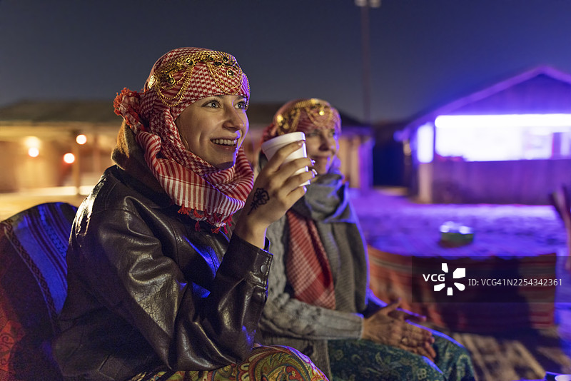 两位戴着头巾的女游客在迪拜沙漠营地享用热饮，同时观看表演。图片素材