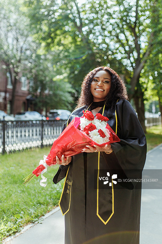 身穿黑色毕业礼服的毕业生手捧花束，在校园户外庆祝成就图片素材