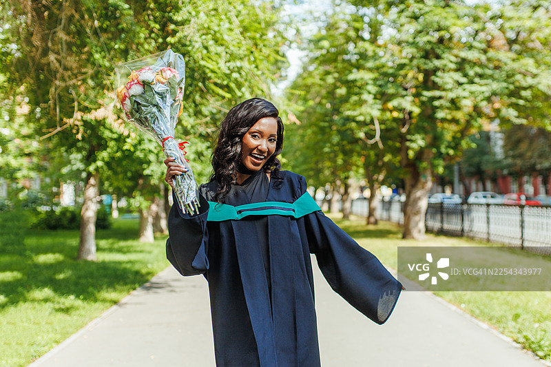 身着毕业礼服、手捧花束的毕业生在阳光明媚的公园小径上庆祝毕业图片素材