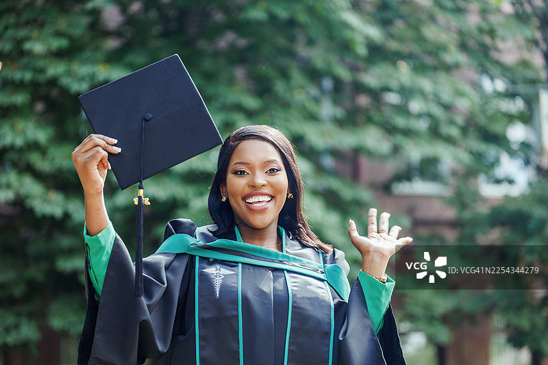 戴着毕业帽和学士服的毕业生在户外庆祝，高举着毕业帽，脸上洋溢着喜悦的笑容图片素材