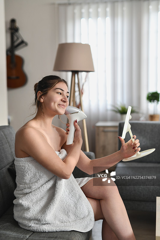 女子裹着浴巾在家中享受面部美容仪和镜子的自我护理水疗时光图片素材