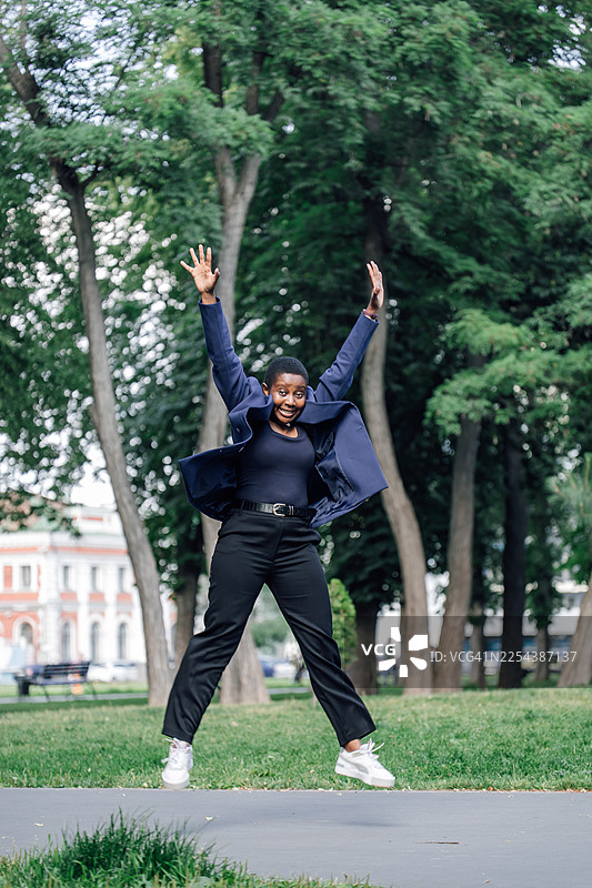 充满活力的女士身穿海军蓝西装外套和黑色裤子，在公园里跳跃，展现出喜悦和自信图片素材