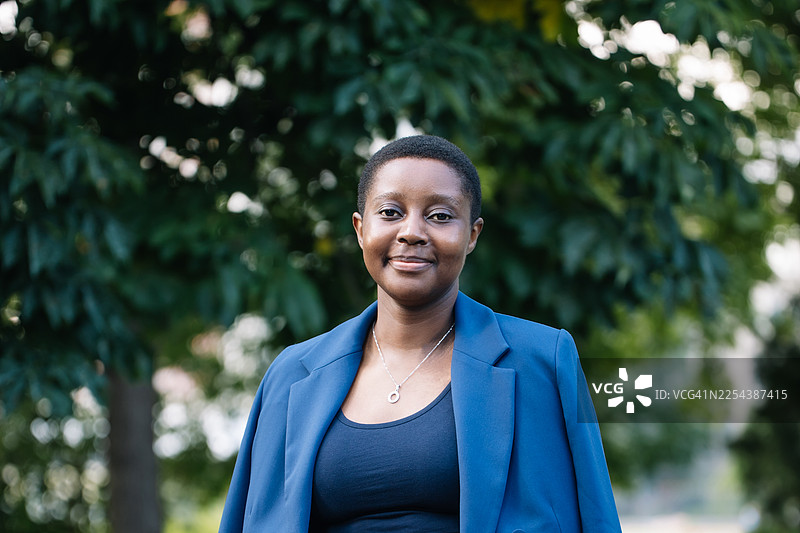 自信的职业女性，身穿蓝色西装外套，面带友善的微笑，站在绿意盎然的公园里图片素材