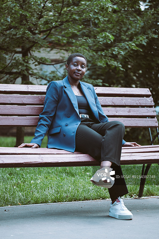 自信的女士身穿蓝色西装外套，悠闲地坐在公园长椅上，拍摄休闲户外肖像图片素材