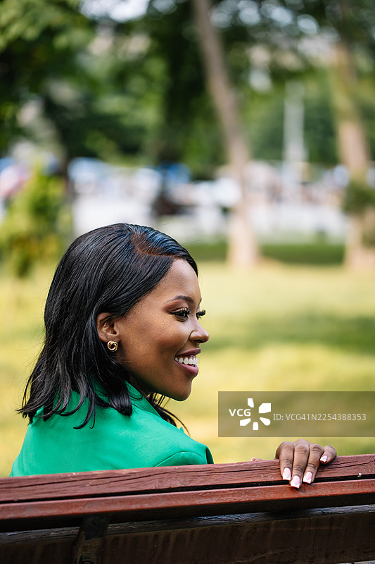 一位身穿绿色西装外套的女士微笑着坐在公园长椅上，享受着阳光明媚的户外时光。图片素材