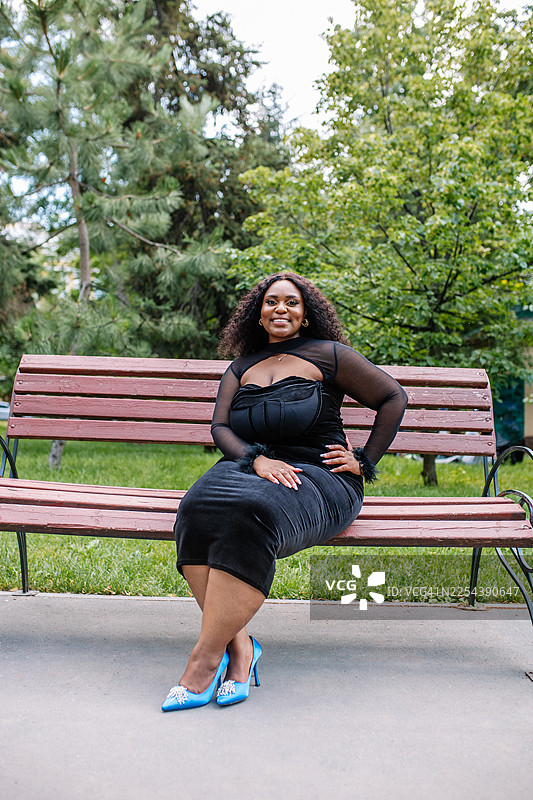 自信的胖女人穿着黑色天鹅绒连衣裙，脚踩蓝色高跟鞋，坐在公园长椅上图片素材