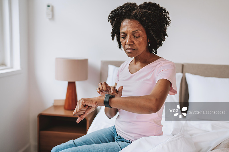 一位年长的黑人女性坐在床边查看智能手表，时间是早晨。图片素材