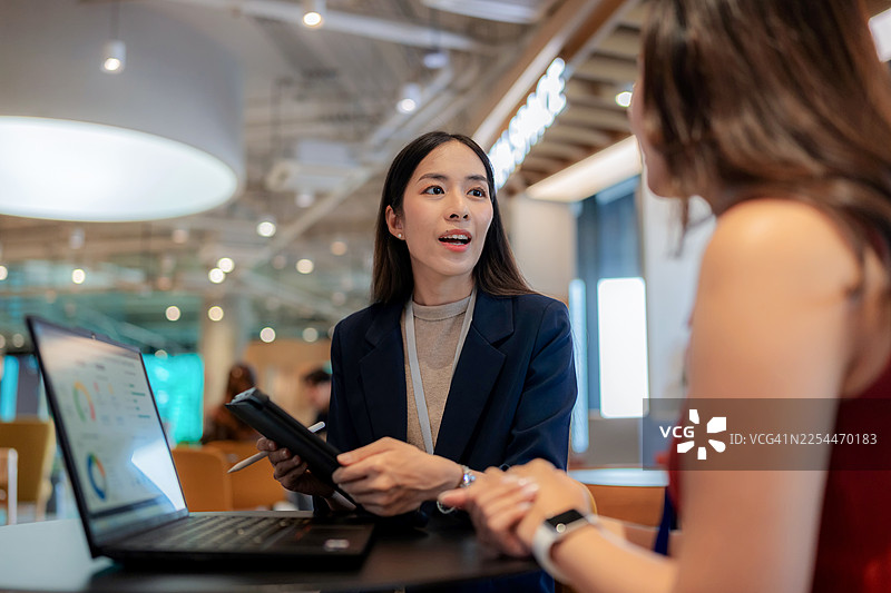 一位专业的亚裔女商人正在现代共享办公空间内，向客户展示笔记本电脑上的财务数据。一位年轻的女企业家在公司会议上讨论项目策略。图片素材