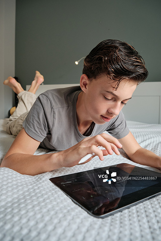 一个十几岁的男孩在床上用触控笔操作数码平板电脑图片素材